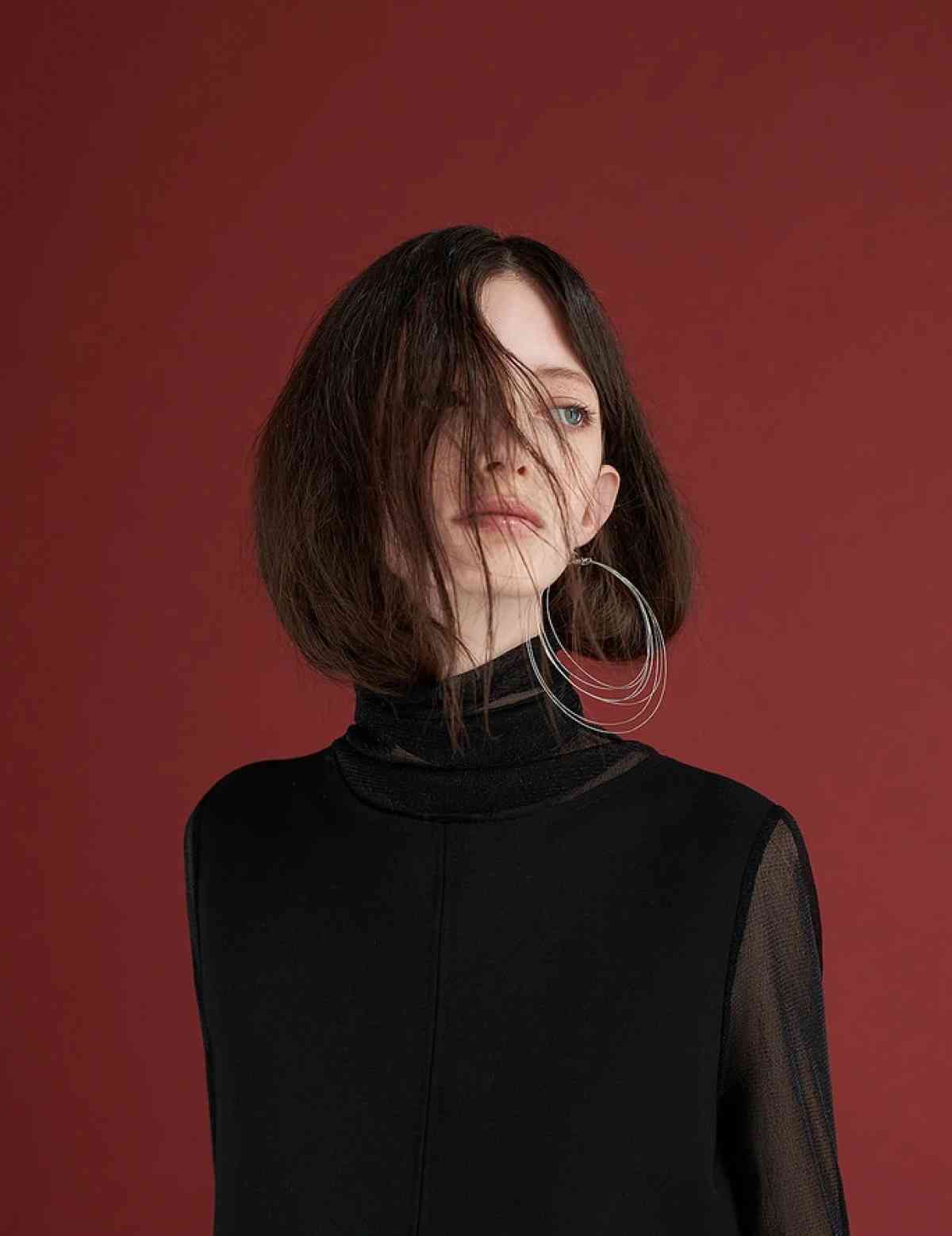 A person with shoulder-length brown hair partly covering their face stands against a red background, embodying the sleek modernity of digital marketing. They are wearing a black turtleneck top and large hoop earrings, effortlessly blending style with the art of SEO.